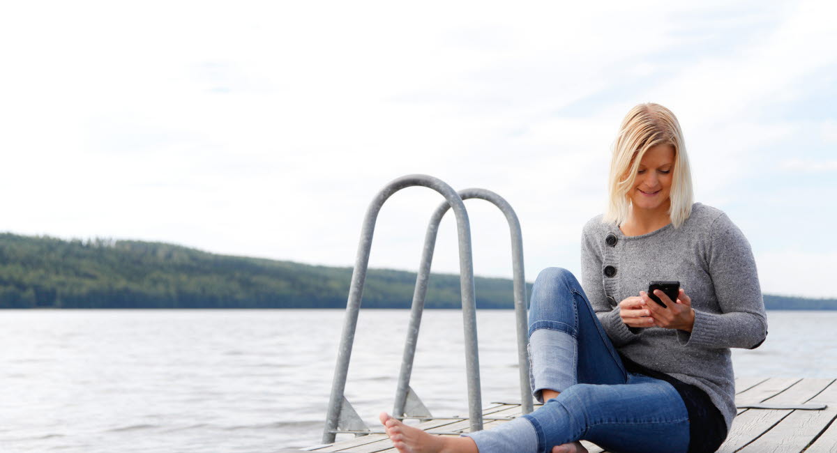 Blond kvinna som sitter på brygga ute i sommarvädret och kollar på sin mobil.